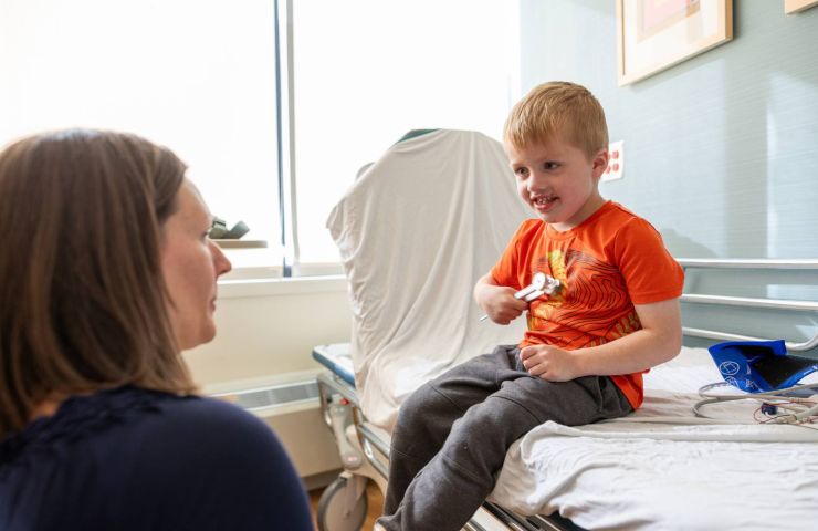 A gillette patient in an orange shirt sits on a hospital bed, looking at a physician and smiling.